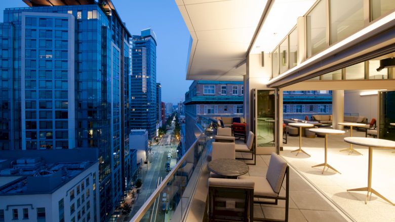 A modern cityscape viewed from a balcony at night, featuring outdoor seating and tall buildings with illuminated windows.