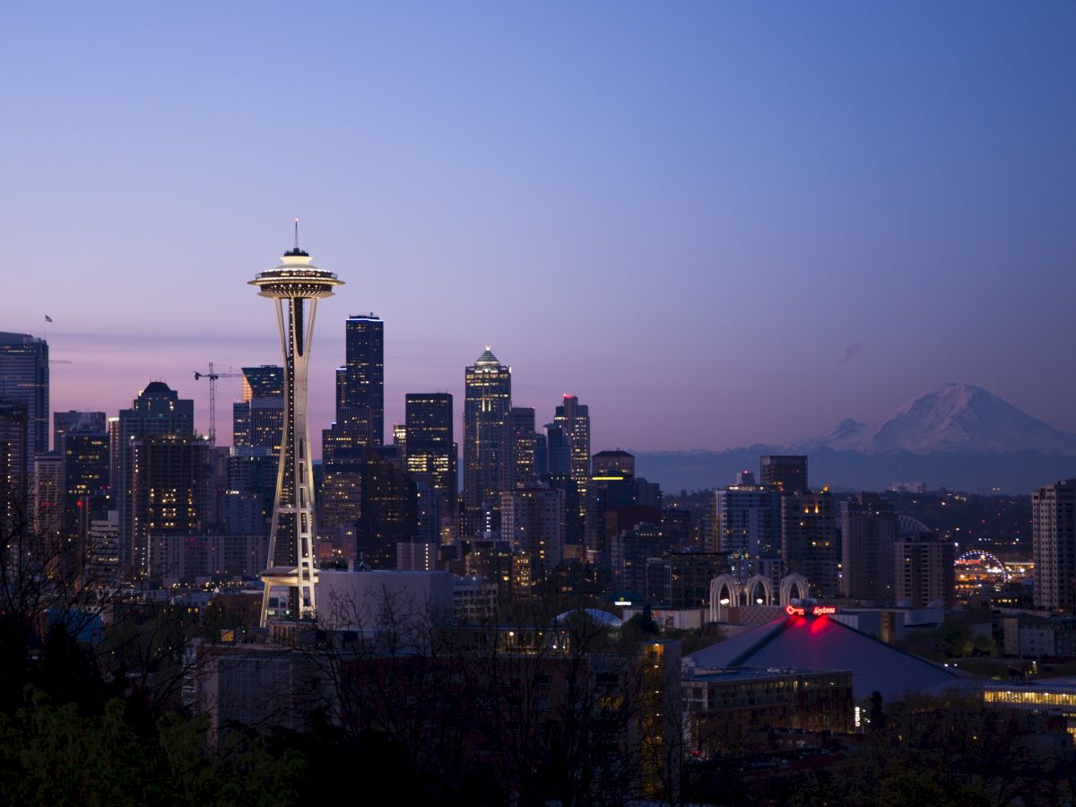 The image shows the Seattle skyline at dusk, featuring the Space Needle and Mount Rainier in the background against a blue and pink sky.
