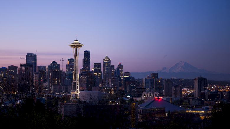 The image shows the Seattle skyline at dusk, featuring the Space Needle and Mount Rainier in the background against a blue and pink sky.