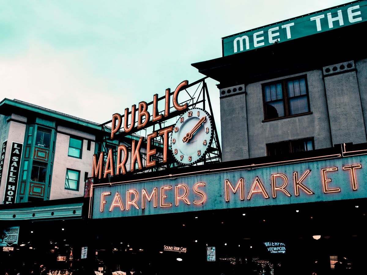 The image shows the iconic sign for a public and farmers market, featuring a clock, against a backdrop of buildings with vintage architecture.