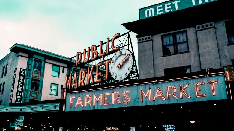 The image shows the iconic sign for a public and farmers market, featuring a clock, against a backdrop of buildings with vintage architecture.