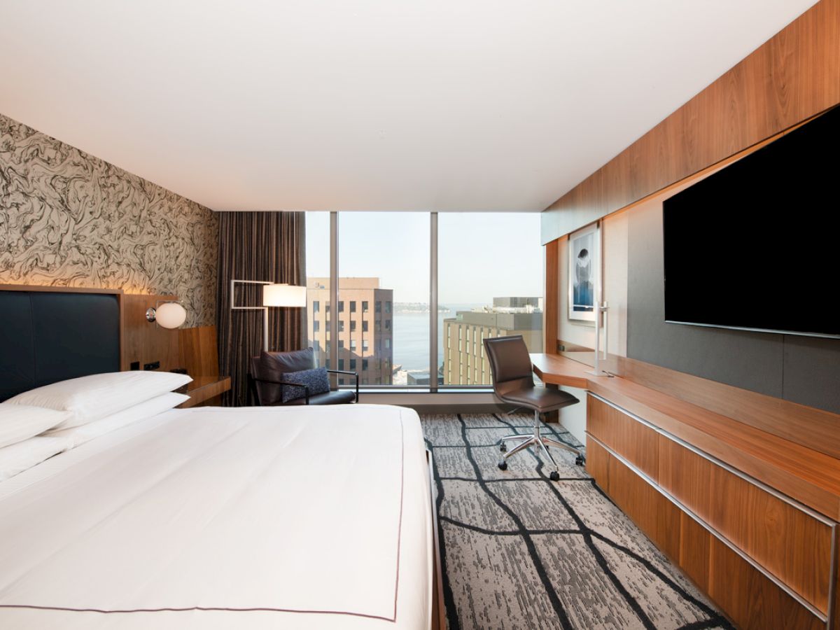 A modern hotel room features a bed, desk, chair, large TV, and a window view of urban buildings and water, with sleek decor.