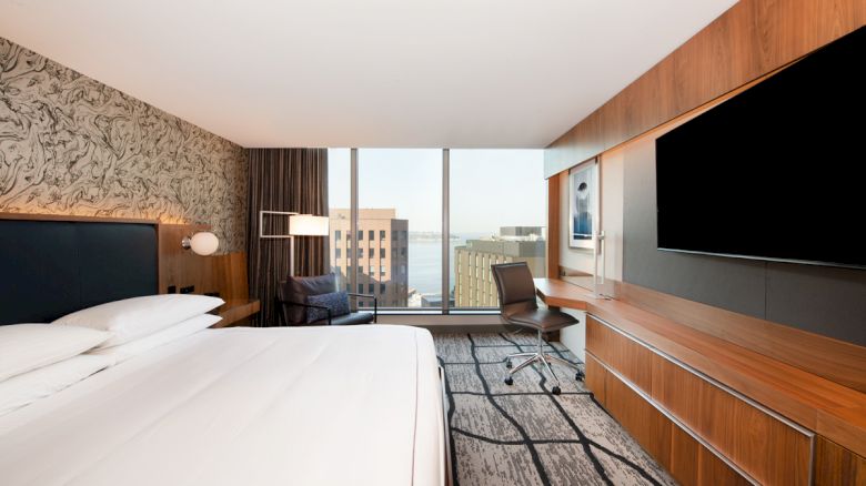 A modern hotel room features a bed, desk, chair, large TV, and a window view of urban buildings and water, with sleek decor.
