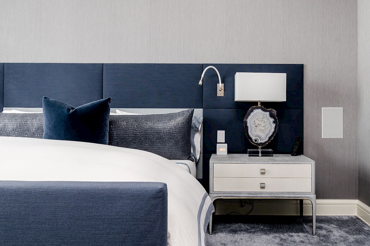 This image shows a modern bedroom with a blue headboard, cozy bedding, a nightstand with a lamp, and a rug on the floor.