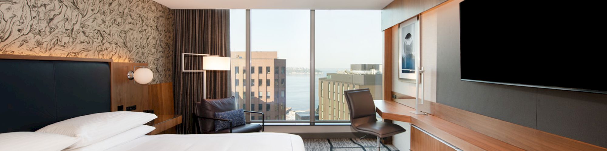 A modern hotel room with a large bed, a desk, a chair, a flat-screen TV, and a window view of city buildings.