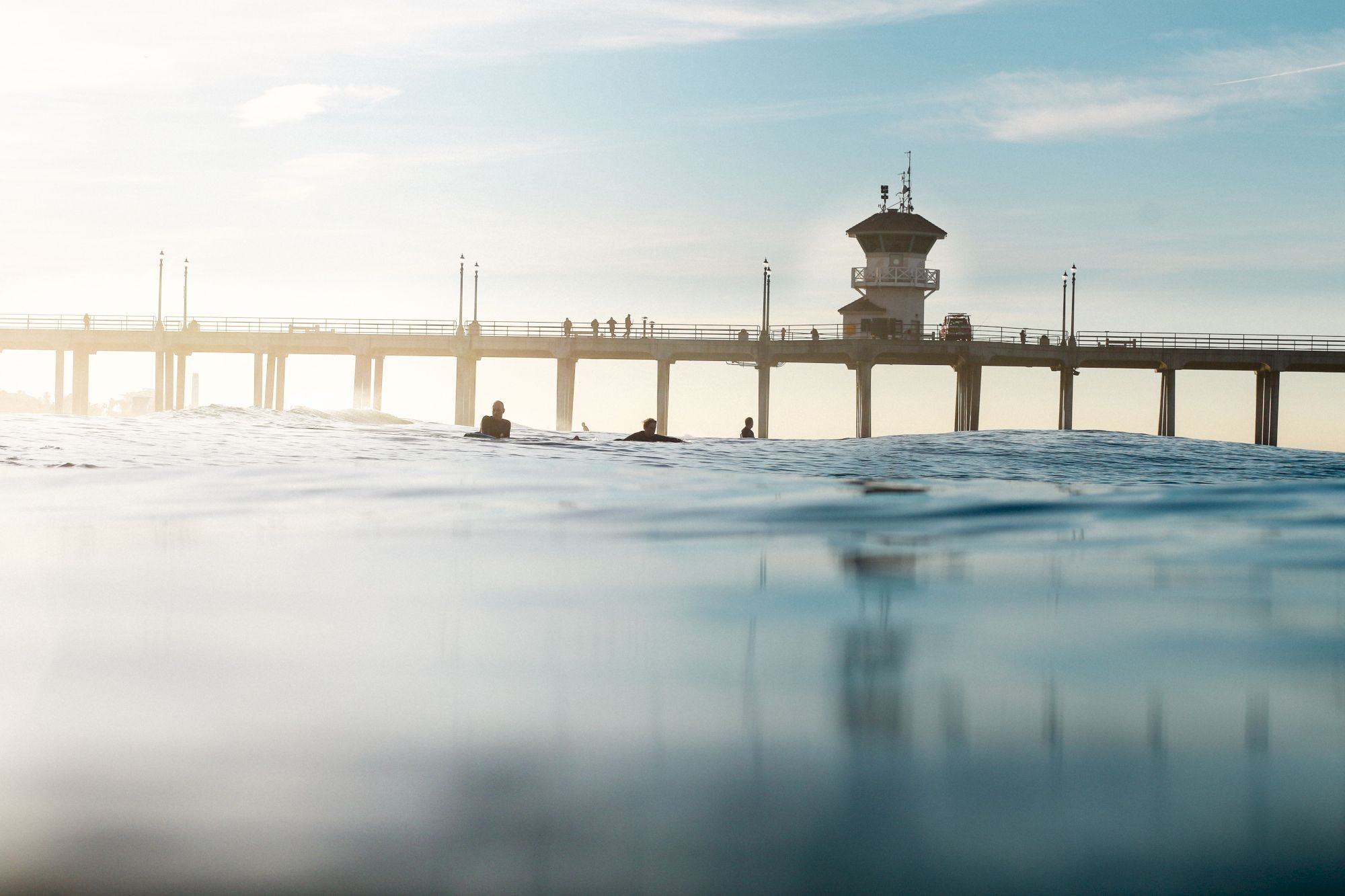 A pier extends over calm water with silhouettes of people standing and surfing as the sun sets, creating a serene coastal scene.